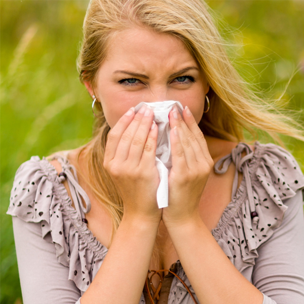 A woman with blonde hair blowing her nose with a tissue outdoors, displaying mild discomfort from allergies possibly eased by rose oil, is set against a blurry green background.