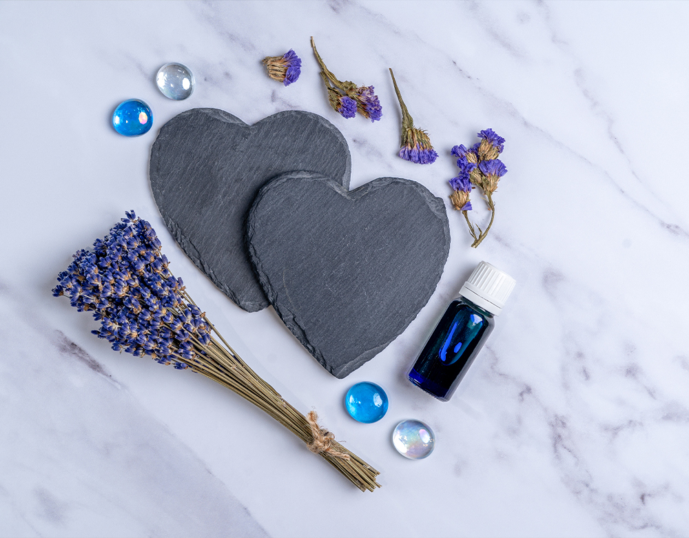 Two slate heart-shaped coasters are accompanied by dried lavender sprigs, glass marbles, and a small bottle of rose oil on a marble background.
