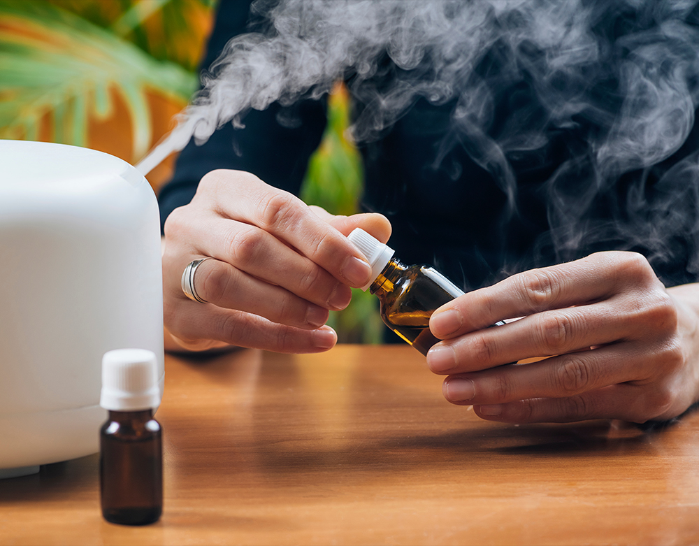 A person is adding essential oil blends to a diffuser, with visible vapor emanating from the device. Two small bottles of essential oil are on the table. A plant is in the background.