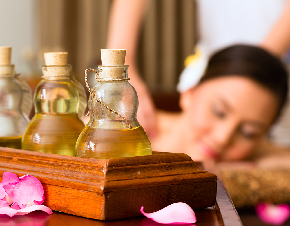 A woman relaxing at a spa with a focus on four bottles of the best essential oils placed on a wooden tray in the foreground, accompanied by scattered pink petals.