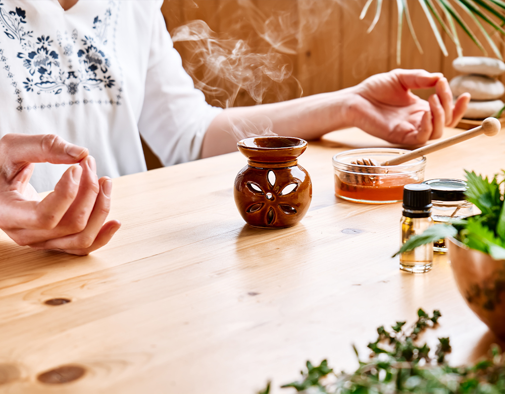 Two people meditating at a wooden table with a steaming ceramic diffuser, essential oil blends, and incense nearby, promoting a calm and relaxing atmosphere.