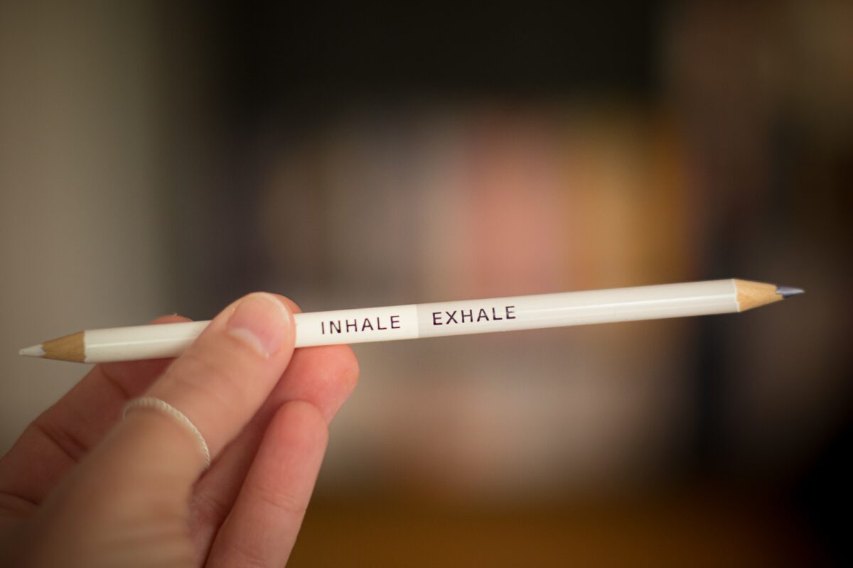 A close-up photo of a hand holding a white pencil with the words "inhale exhale" printed on it. The background, featuring an out-of-focus person, is softly blurred with hints of