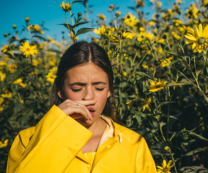 A young woman in a yellow jacket stands amidst tall yellow flowers under a clear blue sky, squinting and touching her nose as if sneezing or reacting to allergies, possibly due to the potent