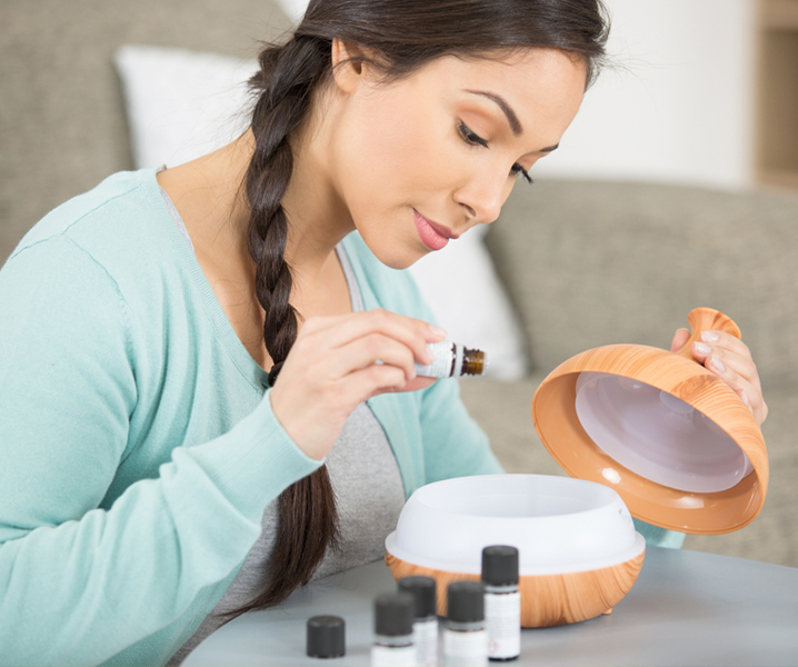 A woman with a long braid adds best essential oils to a diffuser in a cozy living room setting. She appears focused and careful with the bottle.