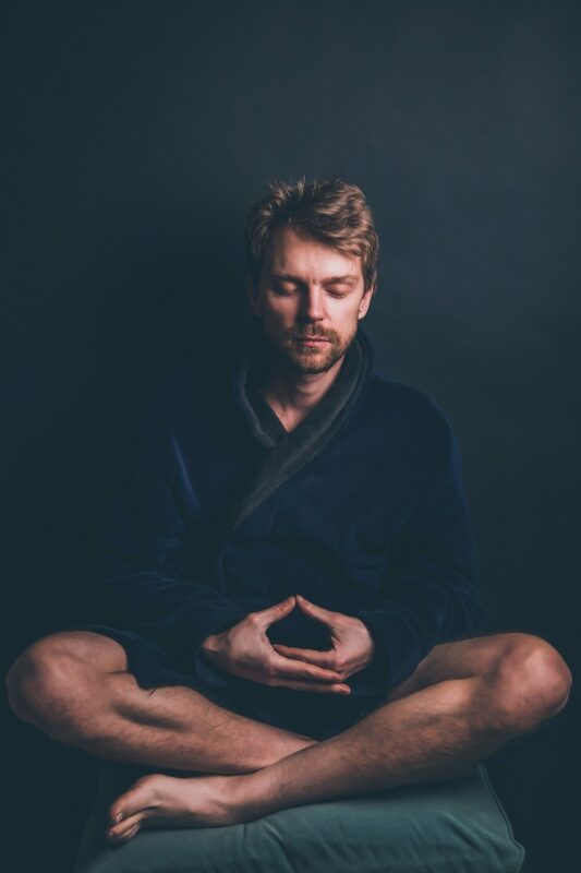 A man in a dark robe meditating with closed eyes, seated cross-legged against a black background. He forms a meditative hand gesture, conveying a sense of calm and concentration, enhanced by the aroma