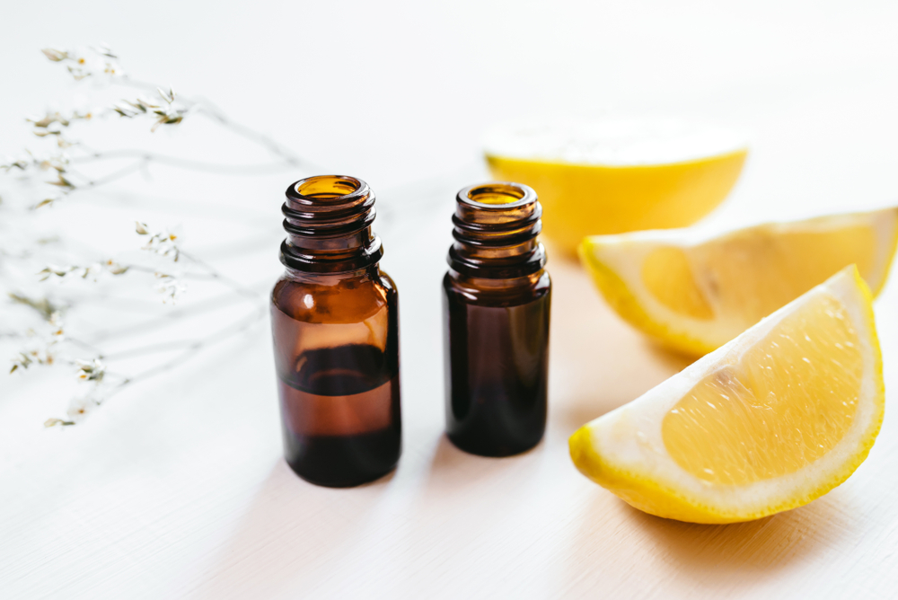 Two small amber glass bottles of the best essential oils next to sliced lemon wedges and white floral sprigs on a bright surface.