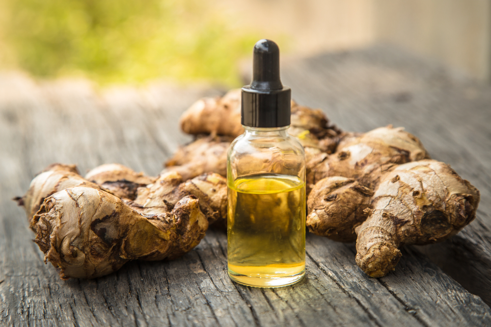 A small glass bottle with a dropper, filled with rose oil, sits in front of a pile of fresh ginger roots on a wooden surface. The focus is on the bottle, with natural light illumin