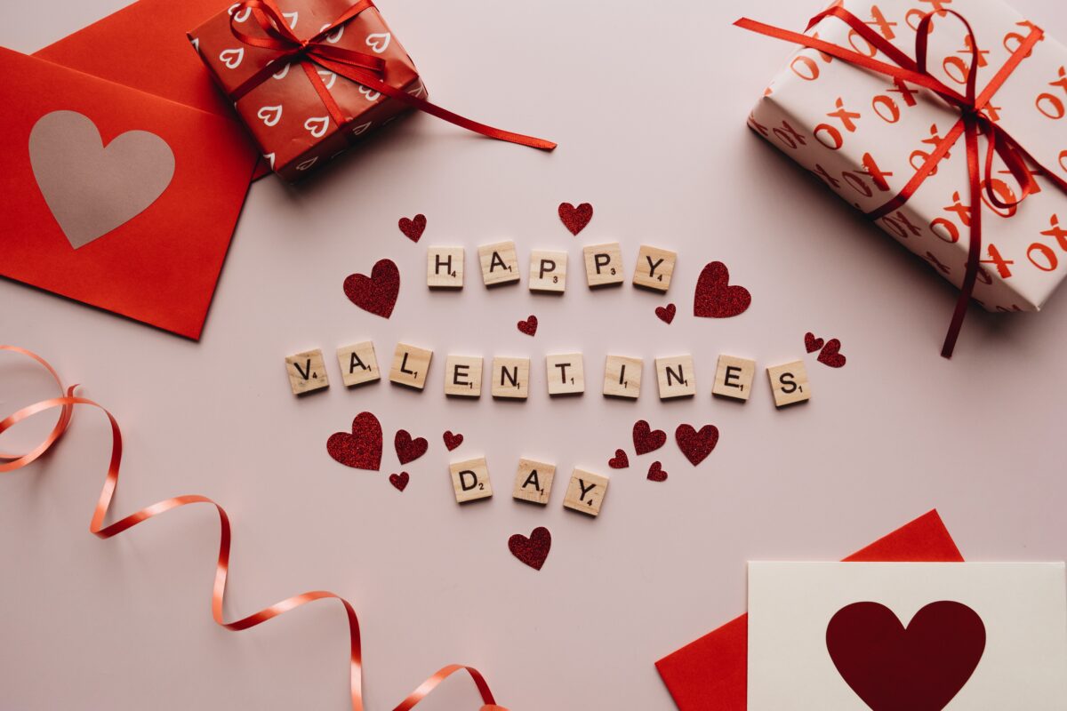 Flat lay of Valentine's Day setting with "happy Valentine's Day" spelled out in letter tiles, surrounded by red gift boxes, heart-shaped decorations, love bird essential oils, and coiled ribbons