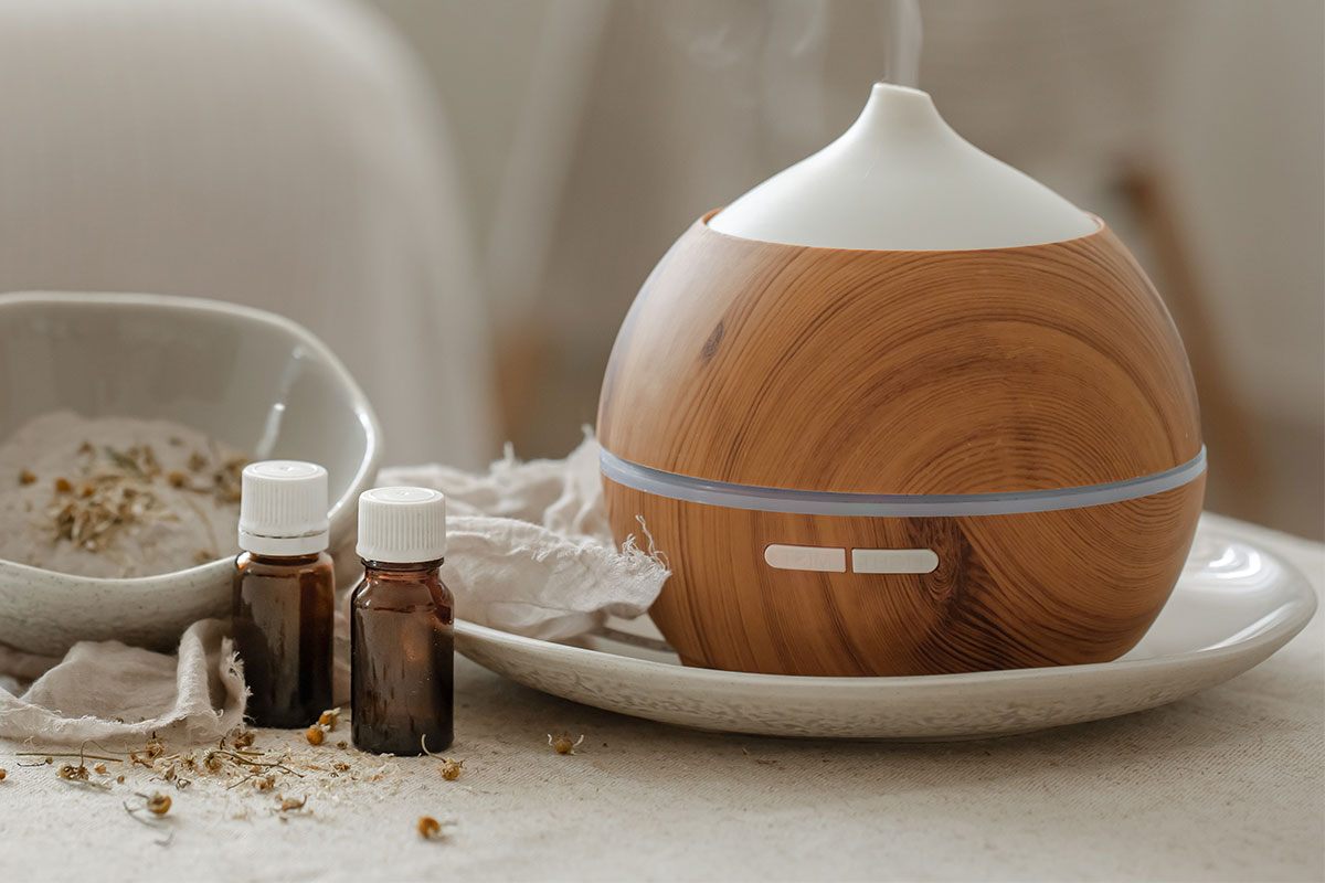 A wooden essential oil diffuser accompanied by two small dark bottles of the best essential oils for cleaning your home and scattered dried herbs, placed on a white ceramic plate in a softly lit setting.