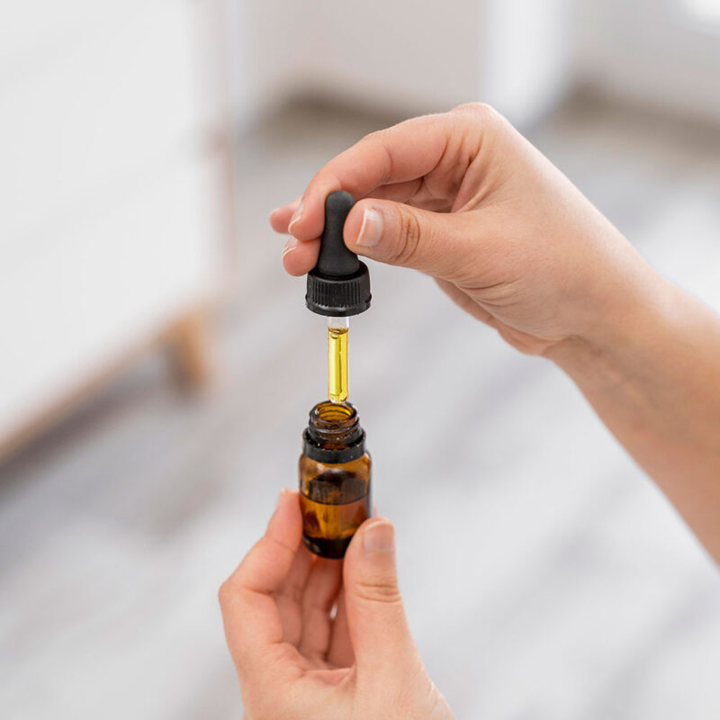 Close-up of hands holding a small amber glass dropper bottle, with one hand squeezing the black dropper to dispense essential oils. A clean, bright interior is softly blurred in the background.