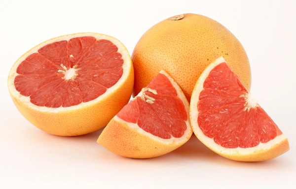 A whole grapefruit alongside a grapefruit cut in half and two wedges, displaying a vibrant pink interior infused with rose oil, arranged on a white background.