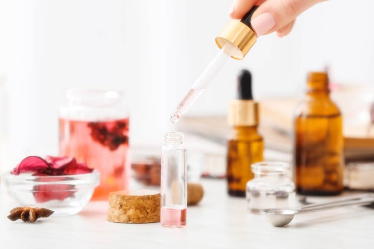 A person using a dropper to incorporate a liquid into a small bottle surrounded by other essential oil bottles, herbs, and a glass jar, set on a wooden table with a white background.