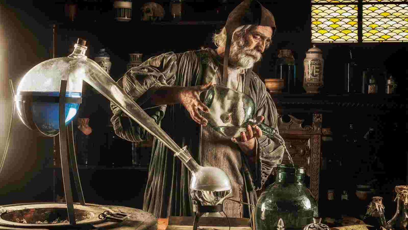 An elderly man dressed in historical clothing works with scientific glassware and equipment in a dimly lit, old-fashioned laboratory setting, focusing on the alchemy of oils.