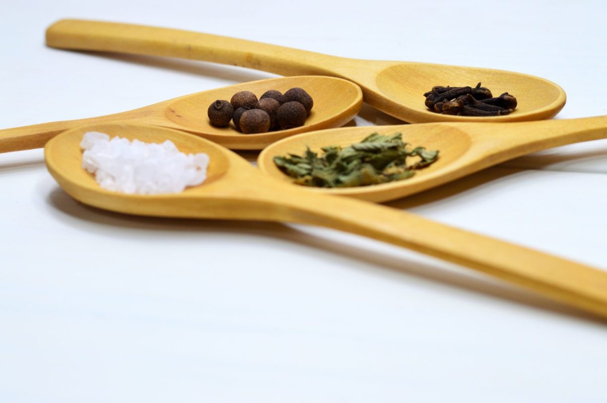 Four wooden spoons on a white surface, each containing Earthsun Essentials spices: coarse salt, whole peppercorns, cloves, and dried herbs. The focus is sharp on the first two spoons