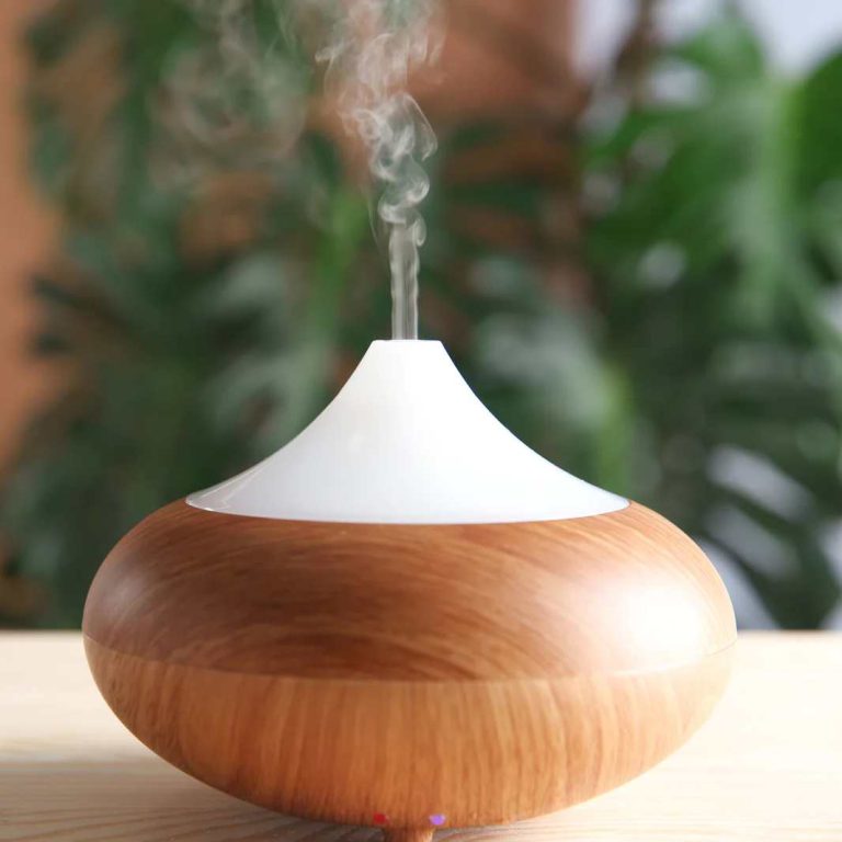 A wooden essential oil diffuser on a table, emitting a thin stream of vapor, with green plants softly blurred in the background. The diffuser has a white top and a rounded wooden base and is
