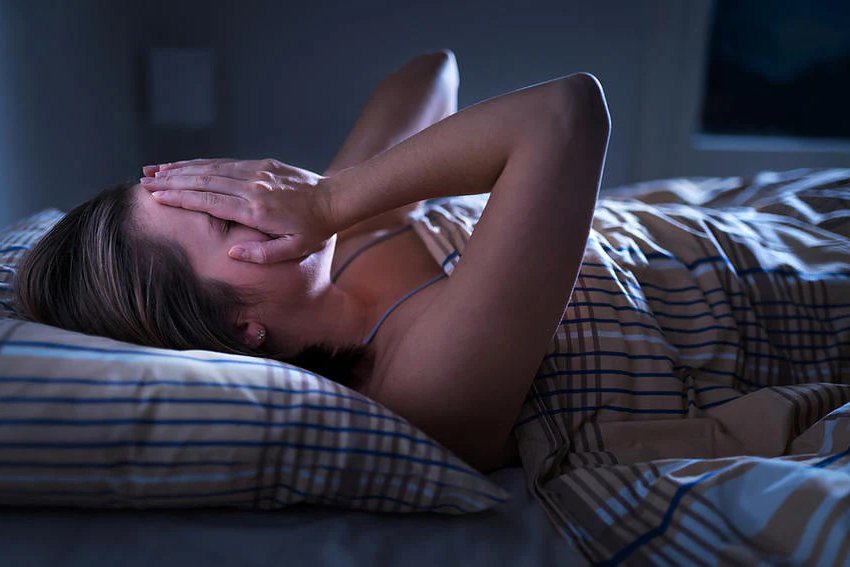 A woman lying in bed, covering her face with her hand, appears distressed or unable to sleep, surrounded by the best essential oils for a good night's rest in a dimly lit room at night