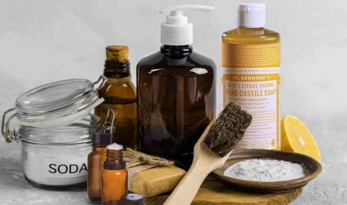 A collection of eco-friendly cleaning supplies including baking soda, essential oil blends, a wooden brush, and liquid soap on a marble surface.