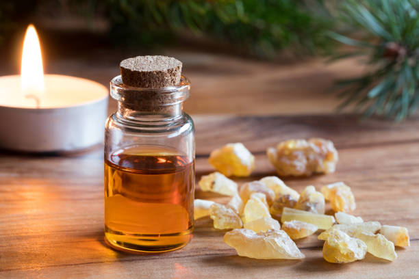 A small glass bottle with a cork stopper filled with amber liquid, next to chunks of frankincense resin and a lit candle, with pine branches in the background. Auto Draft